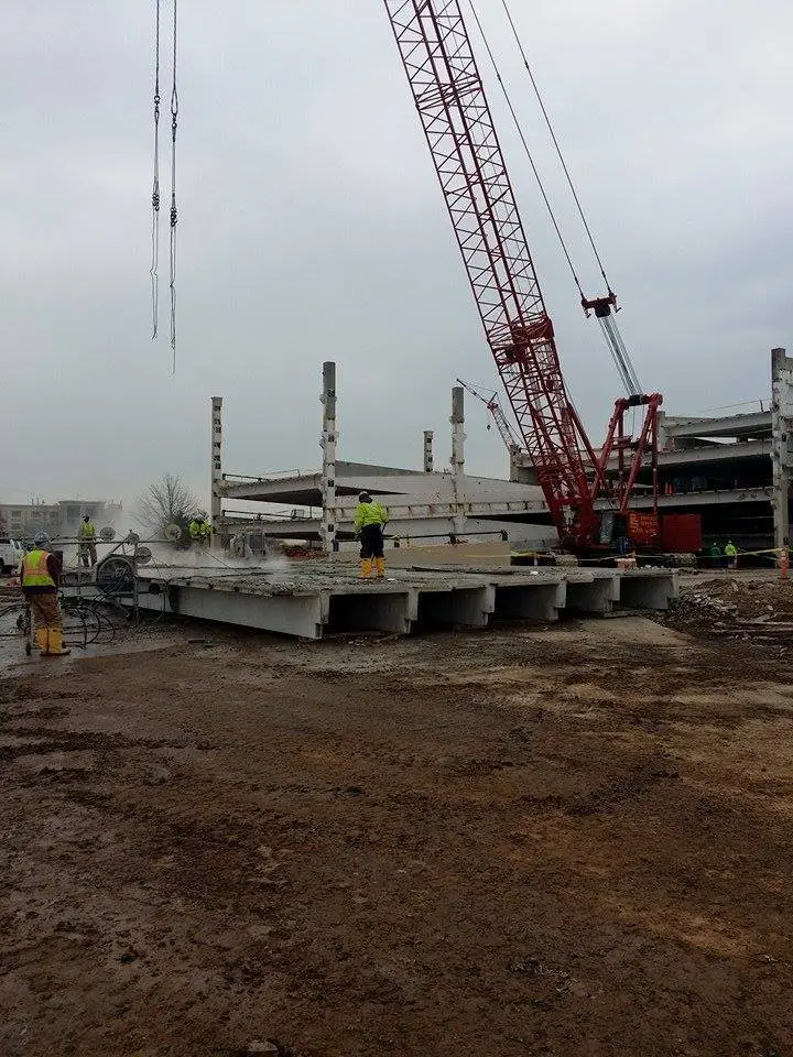 Image of a worksite. Showcasing men in neon work vests, a large concrete structure and a crane