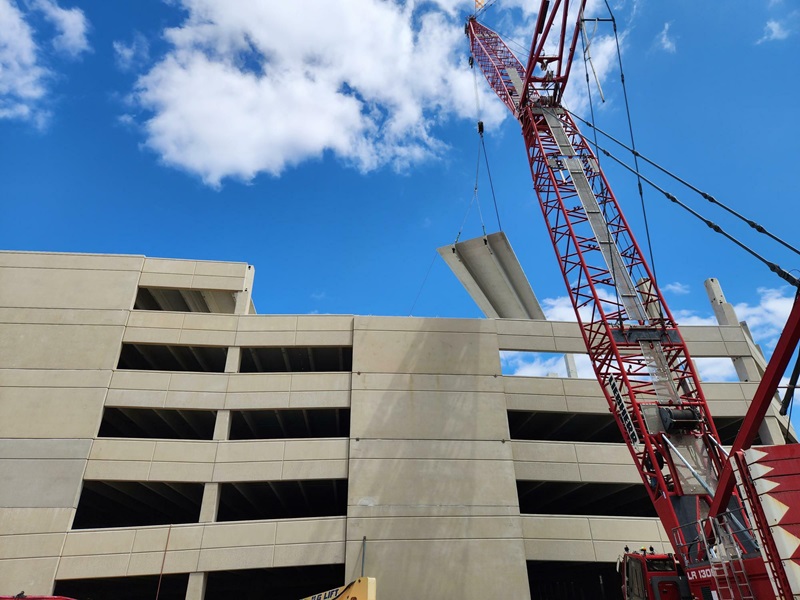 A red crane lifting up a rectangular piece above a parking garage structure.